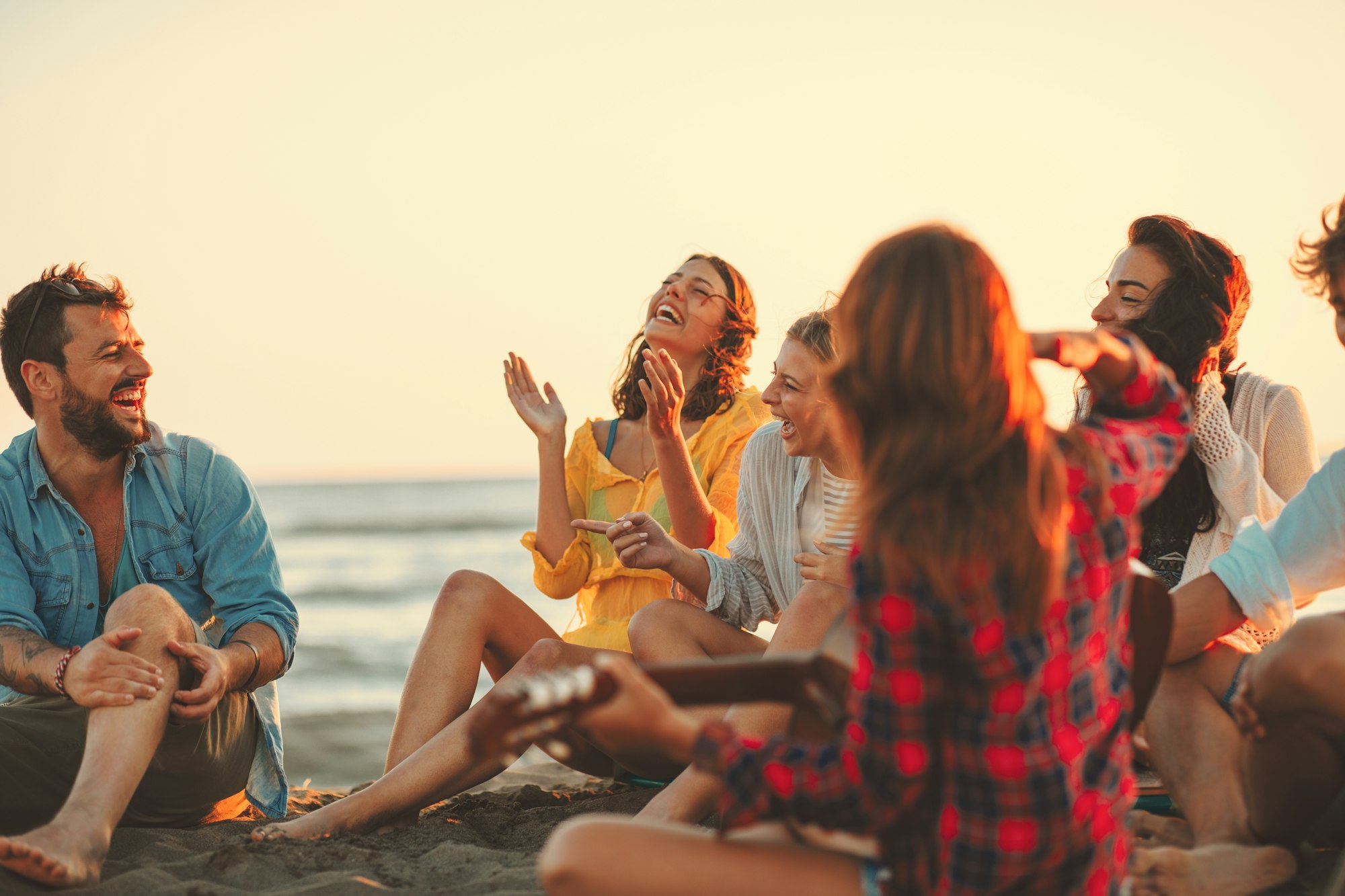 Happy friends sitting on the beach singing and playing guitar during the sunset